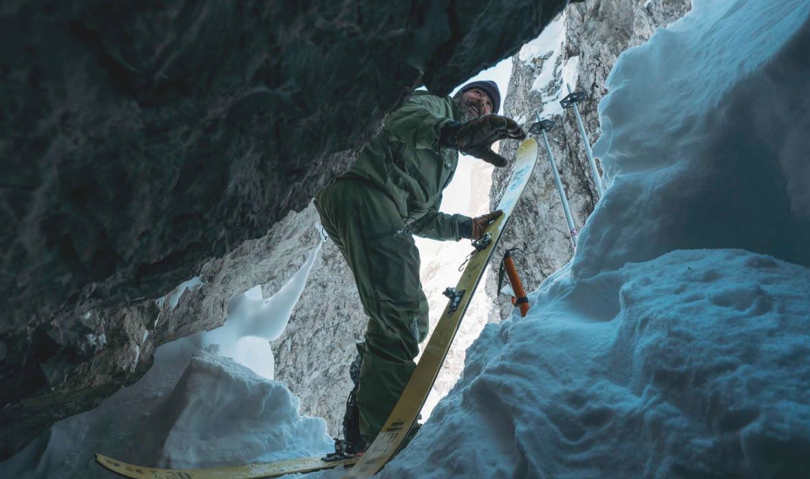 Ein Skifahrer in einer engen, schneebedeckten Felsspalte beim Justieren seiner Skiausrüstung; er trägt grüne Outdoor-Kleidung und ist von Schnee und steilen Felsen umgeben.